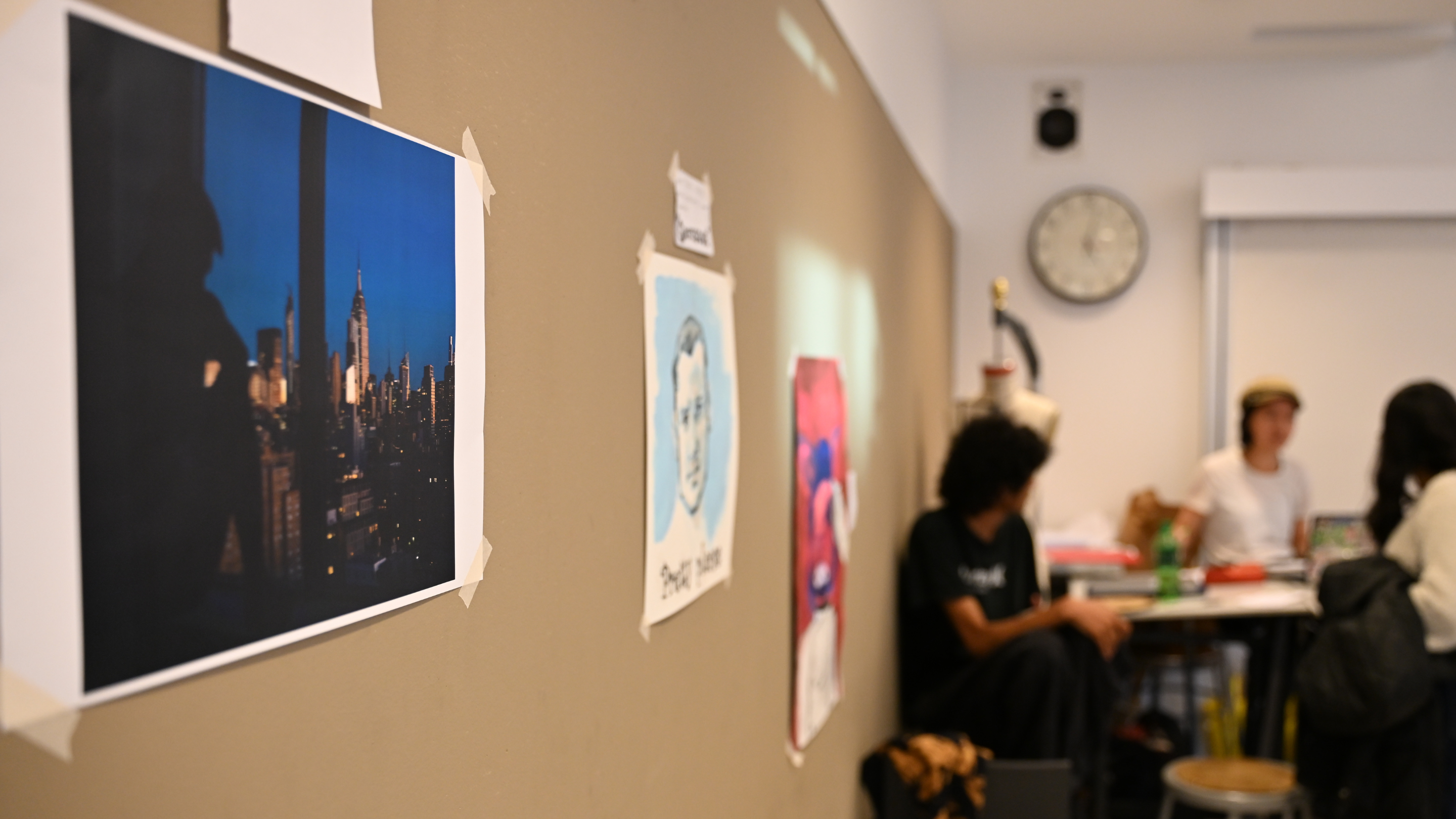 Photo of a classroom wall with taped-up art prints and photos, including a cityscape of New York. In the background, three students are gathered around a table in discussion.