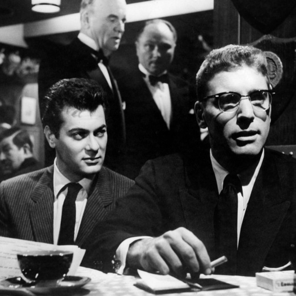 Black and white screencap of film. Tony Curtis and Burt Lancaster sit at a table in a dimly lit restaurant/club. 