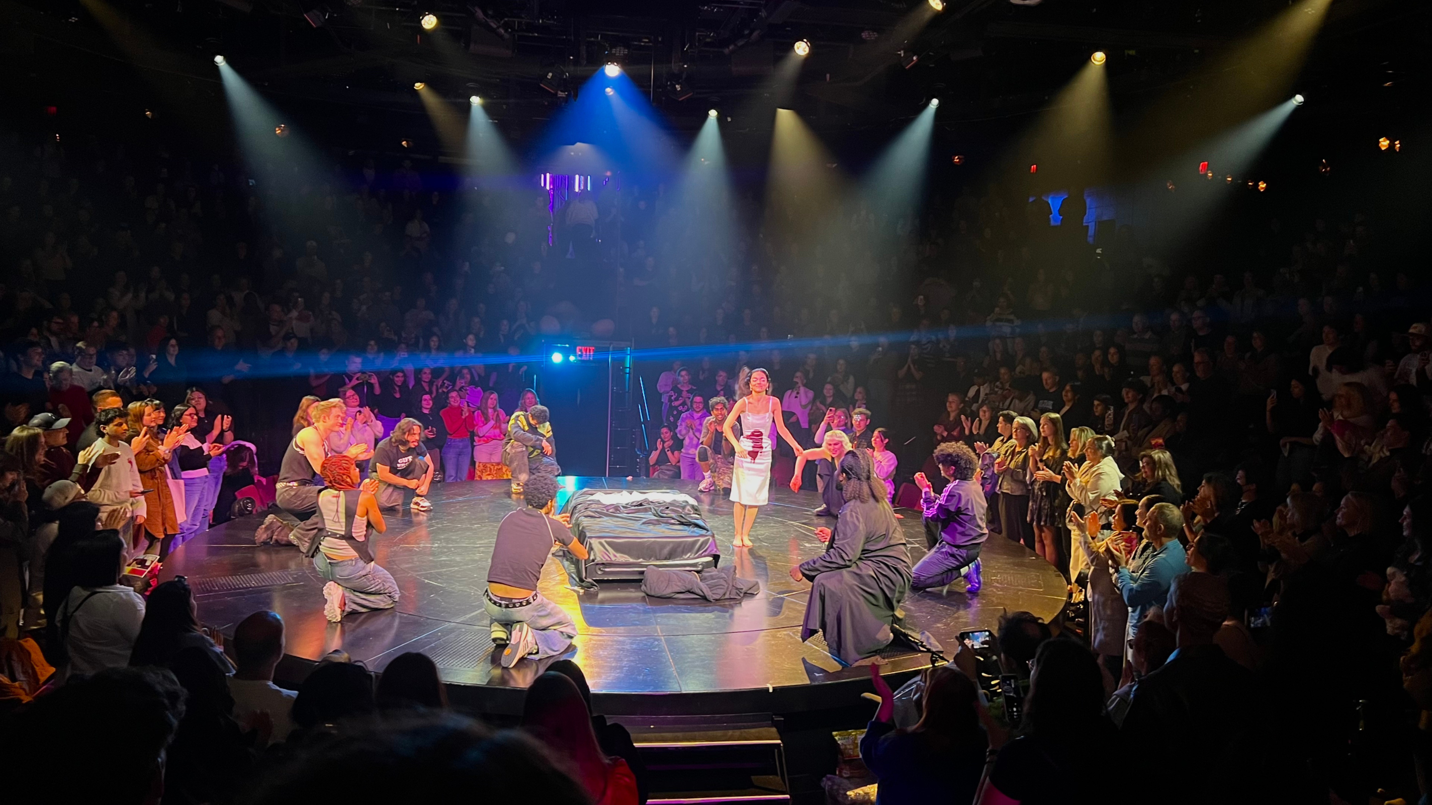 Actors gathered around a dimly lit circular stage during bows, surrounded by an audience in a standing ovation.