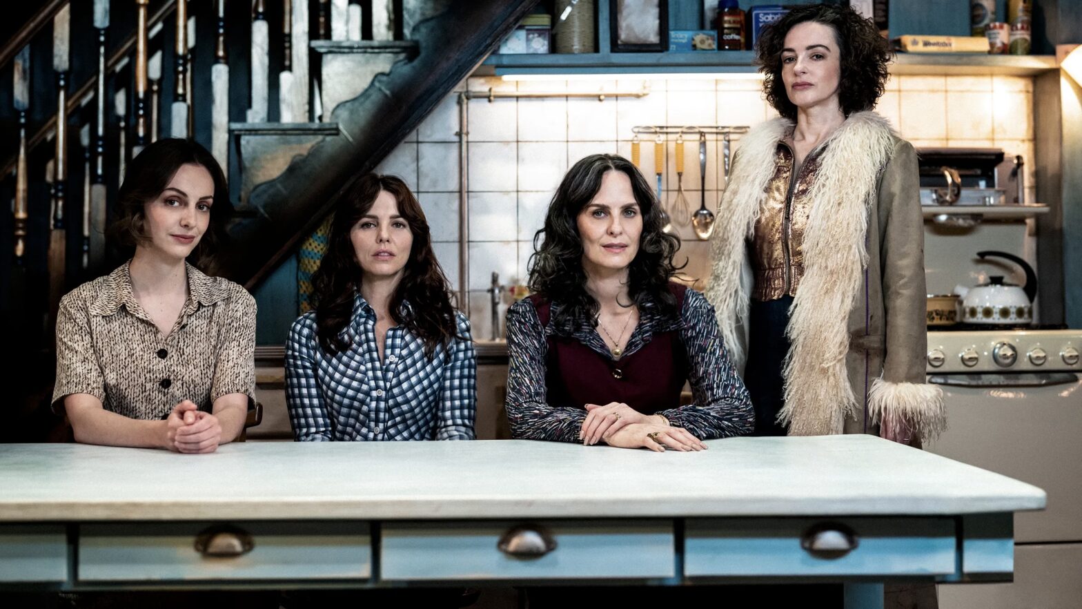 Four sisters around a kitchen table in a rustic setting with a staircase and tiled wall in the background.