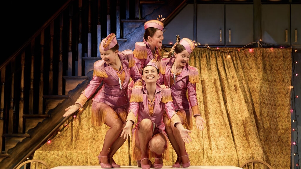 Four performers in pink costumes pose energetically on stage with a gold curtain backdrop and stairs on the left.