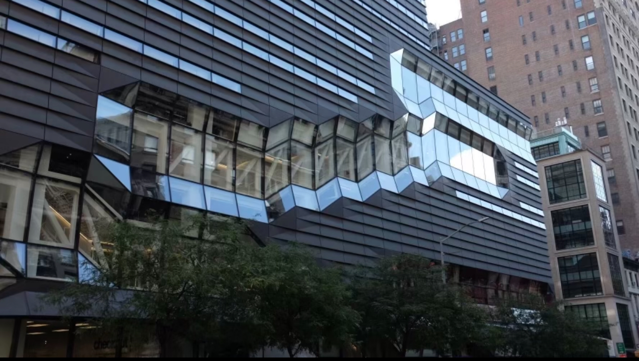 An image of the University Center, a large building with horizontal lines of steel and glass windows. To the right, a tall brown brick building stands.