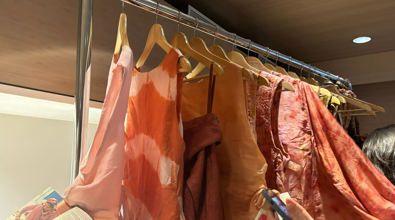 A collection of warm-toned garments,, made by Saheli Women,, hang from a rack. Attendees touching and taking photos of the clothes.