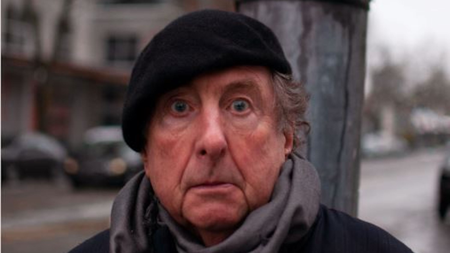 Eric Idle, looking into the camera outside in a suburban neighborhood.