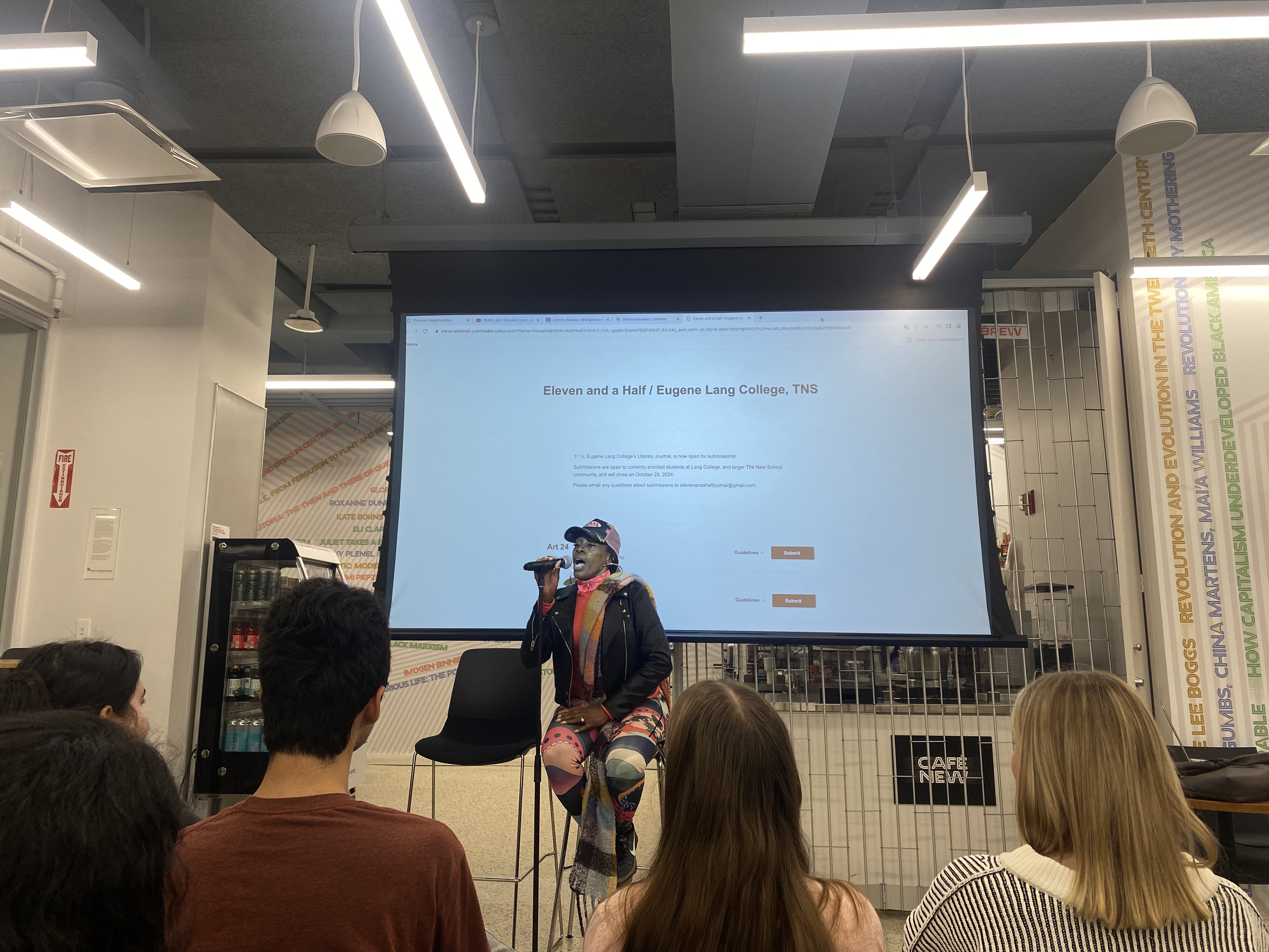 A woman is sitting on a chair holding the microphone in her hand in front of her mouth with the backdrop reading “Eleven and a Half” as students watch