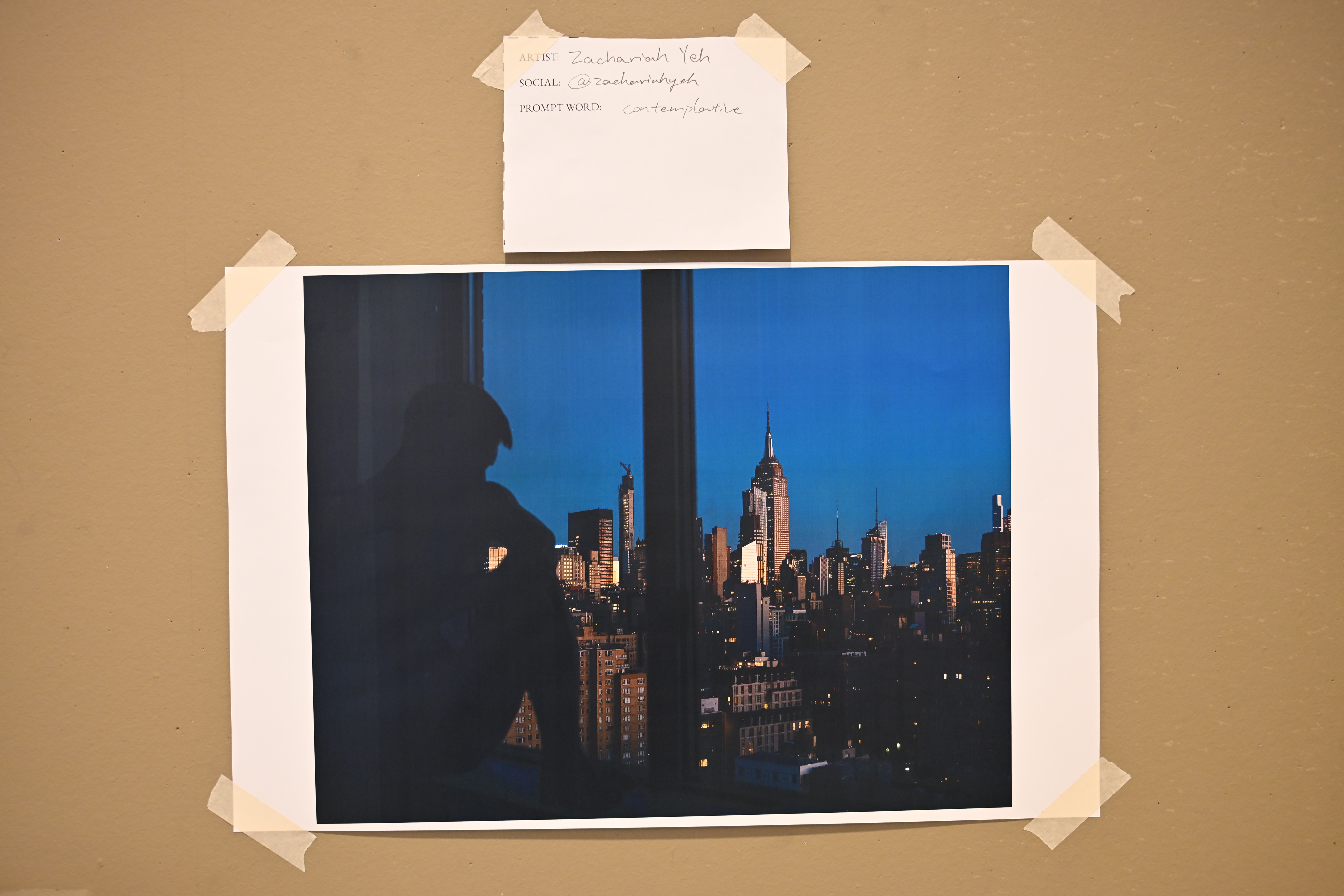 Printed photograph taped to the wall, depicting the NYC skyline and a shadow figure sitting facing a window.