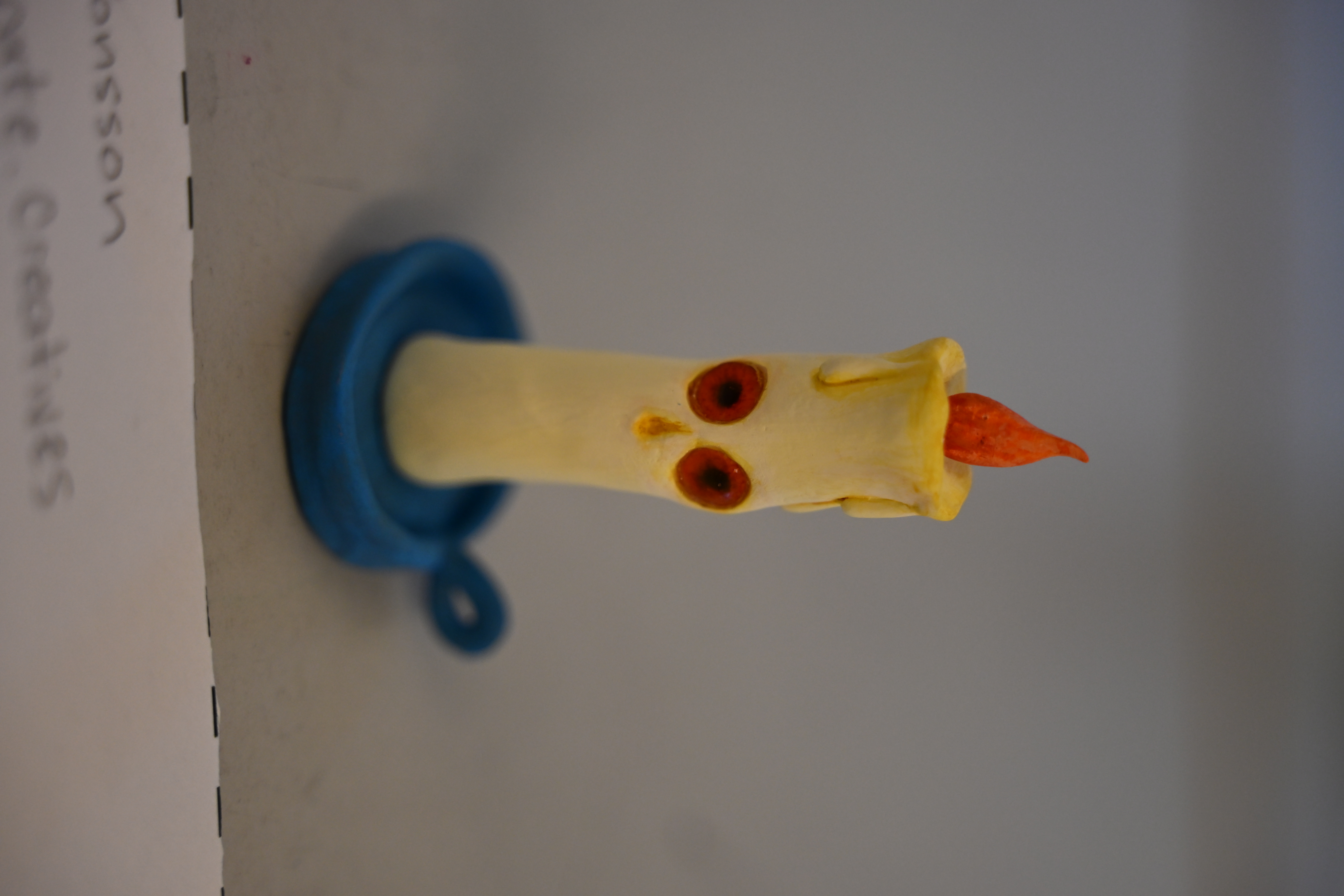 Clay mini sculpture of a flame ignited candle, with wax dripping towards its eyes and face, on top of a blue candle holder.