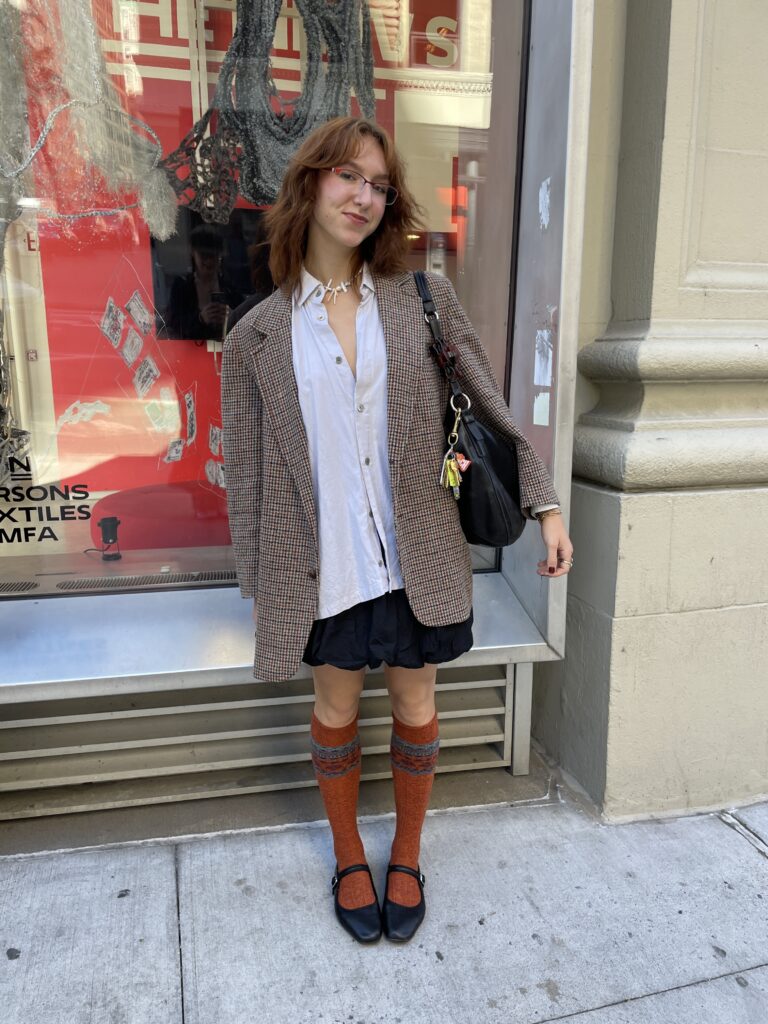 Student stands in front of the Parsons 13th Street building wearing an orange gingham oversized blazer on top of a loose white button-down shirt, a black bubble skirt, with burnt orange knee-high socks, and black Mary Janes. 
