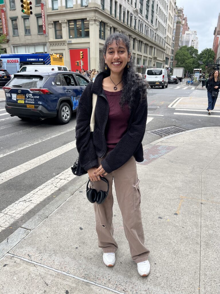 Student wears white sneakers, tan cargo pants, a tote bag, a fuzzy black jacket, a burgundy top, gold earrings, a brown necklace, and two rings on each of her hands. 