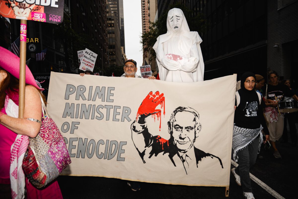 Demonstraters holding up sign that reads "Prime Minister of Genocide" with a posed Benjamin Netanyahu, holding his hand up which is covered in blood