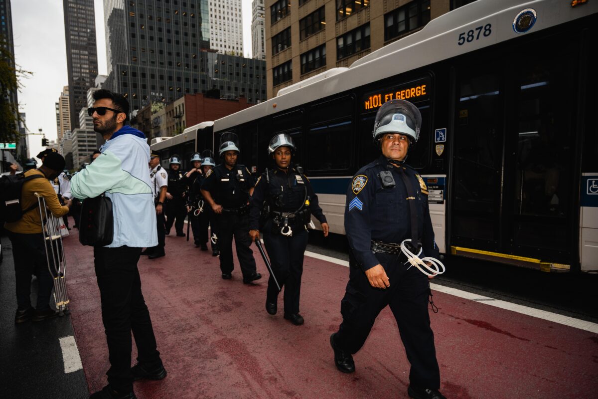 Police officers walking down the street in a single filed line, with zip ties and batons, and in riot gear.