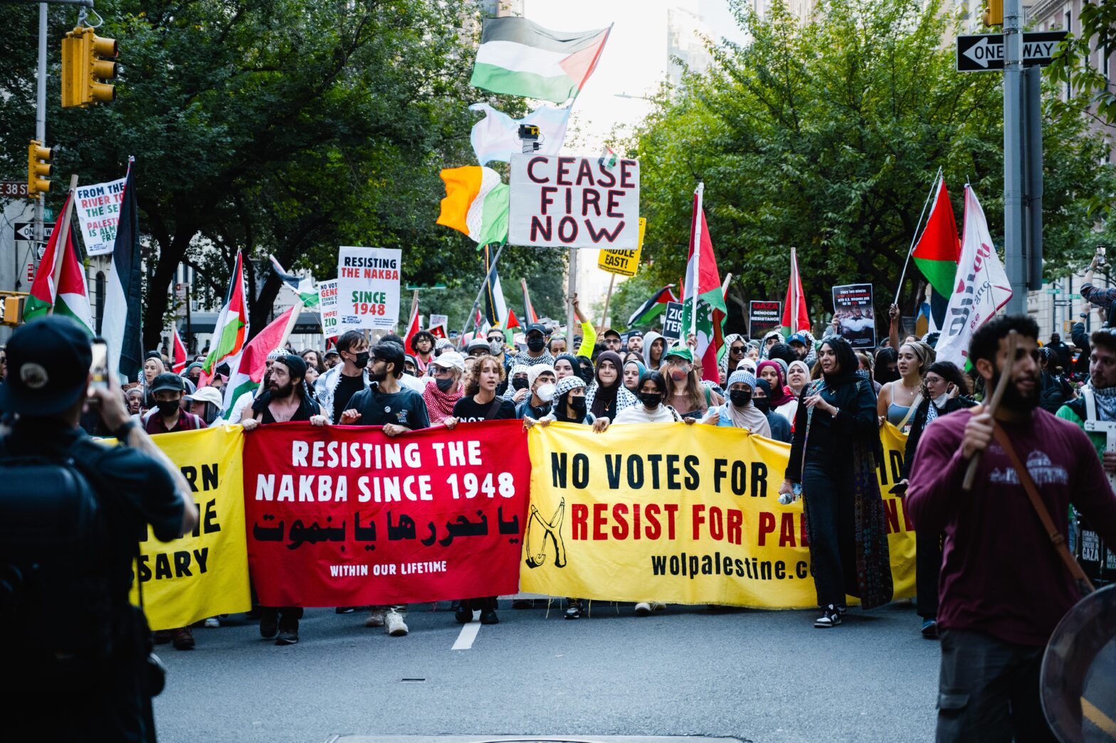 A group of hundreds of protestors, primarily wearing white, hold three banners, which alternate between yellow, red, and yellow from left to right. In white letters, the middle banner reads “resisting the nakba since 1948.” Demonstrators hold signs that read “ceasefire now” and Palestinian flags.