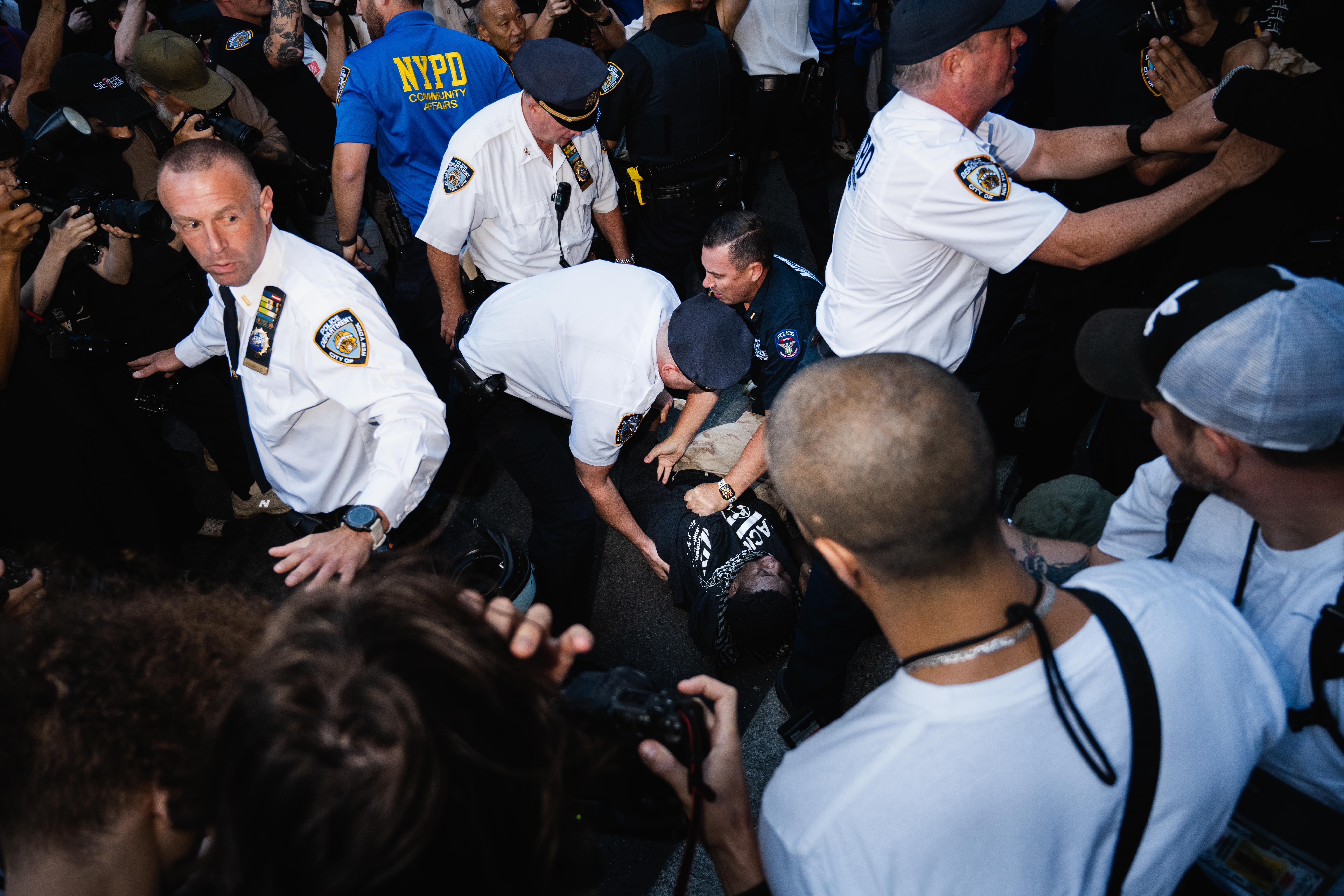 Four NYPD officers dressed in white, two in black, and one Community Affairs officer in blue surround a demonstrator on the ground. Two of the officers have their hands on the demonstrator, and other demonstrators crowd around the officers. 