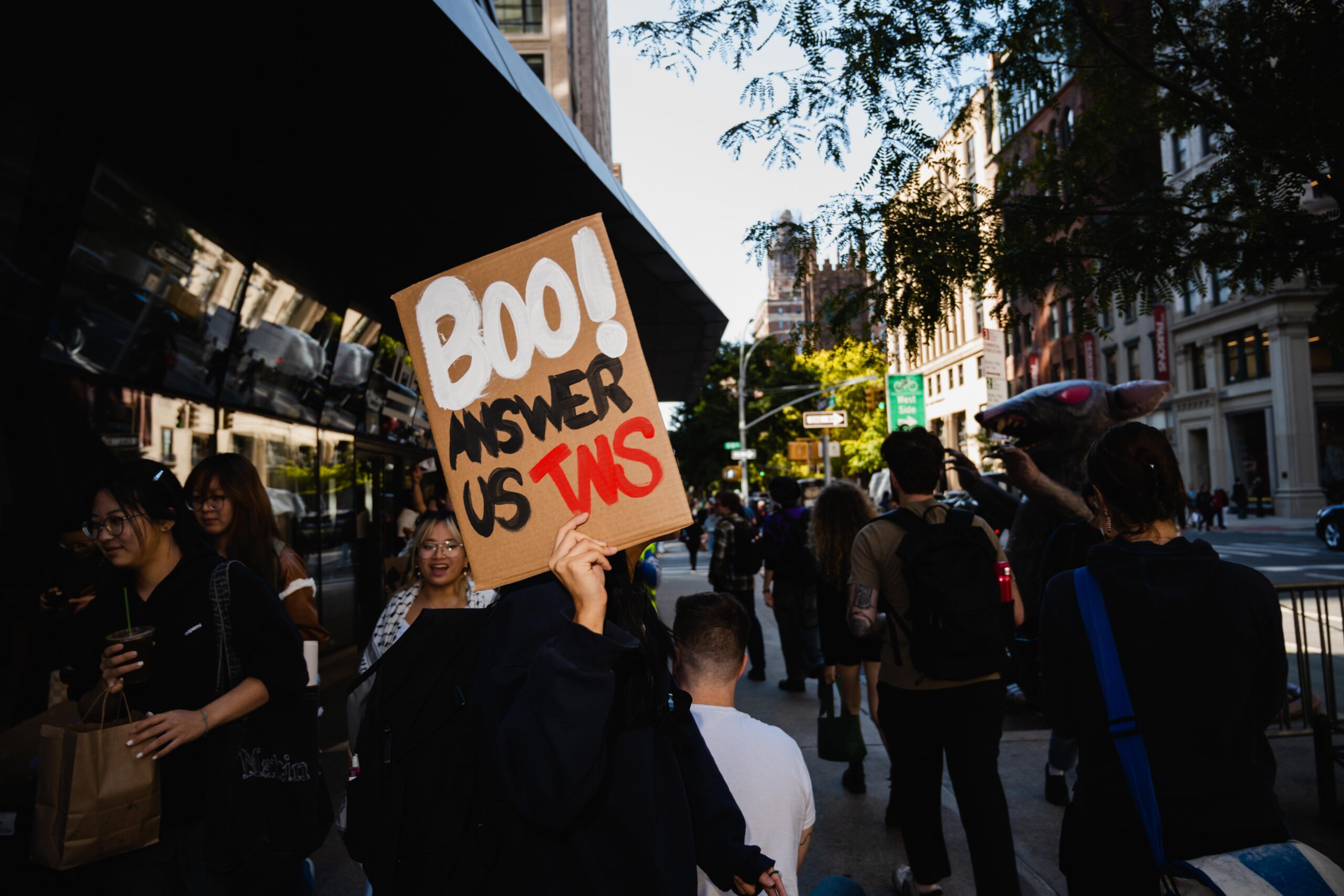 Protesters holding up a sign saying, “Boo! Answer us TNS”, while other protesters walk in the background.