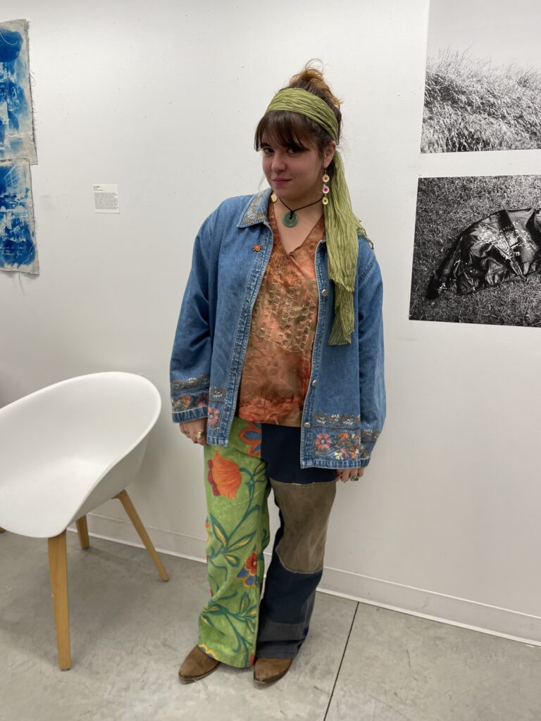 Student stands inside the Parsons 13th Street Building wearing handmade patchwork pants with hand painted green and floral accents, a tye-dye orange and brown shirt with stitching details, a denim jacket with floral embroidery on the edges, triple-decker eye earrings, a circular jade necklace, and a green scarf as a headband. 
