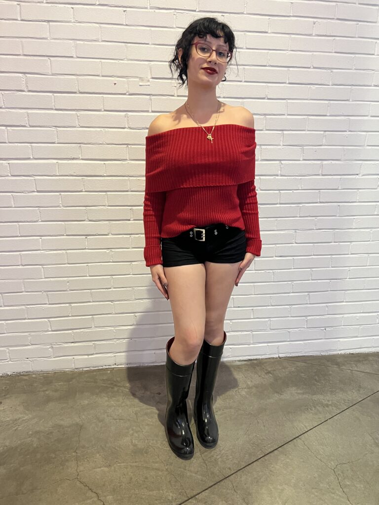 Student wears a red sweater top,  black shorts held up by a large belt, gold jewelry, red cat-eye glasses, and black calf-length boots.