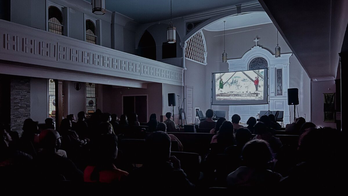 Image of a crowd watching a film projected on a large screen in a dimly lit room with high ceilings and arched windows.