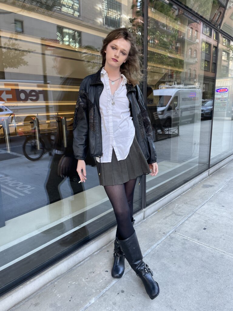 Student stands outside of the University Center building smoking a cigarette. She is wearing a fitted black leather jacket, a ruffled white button-down, a gray pleated mini skirt, black sheer tights, and black buckle boots. 
