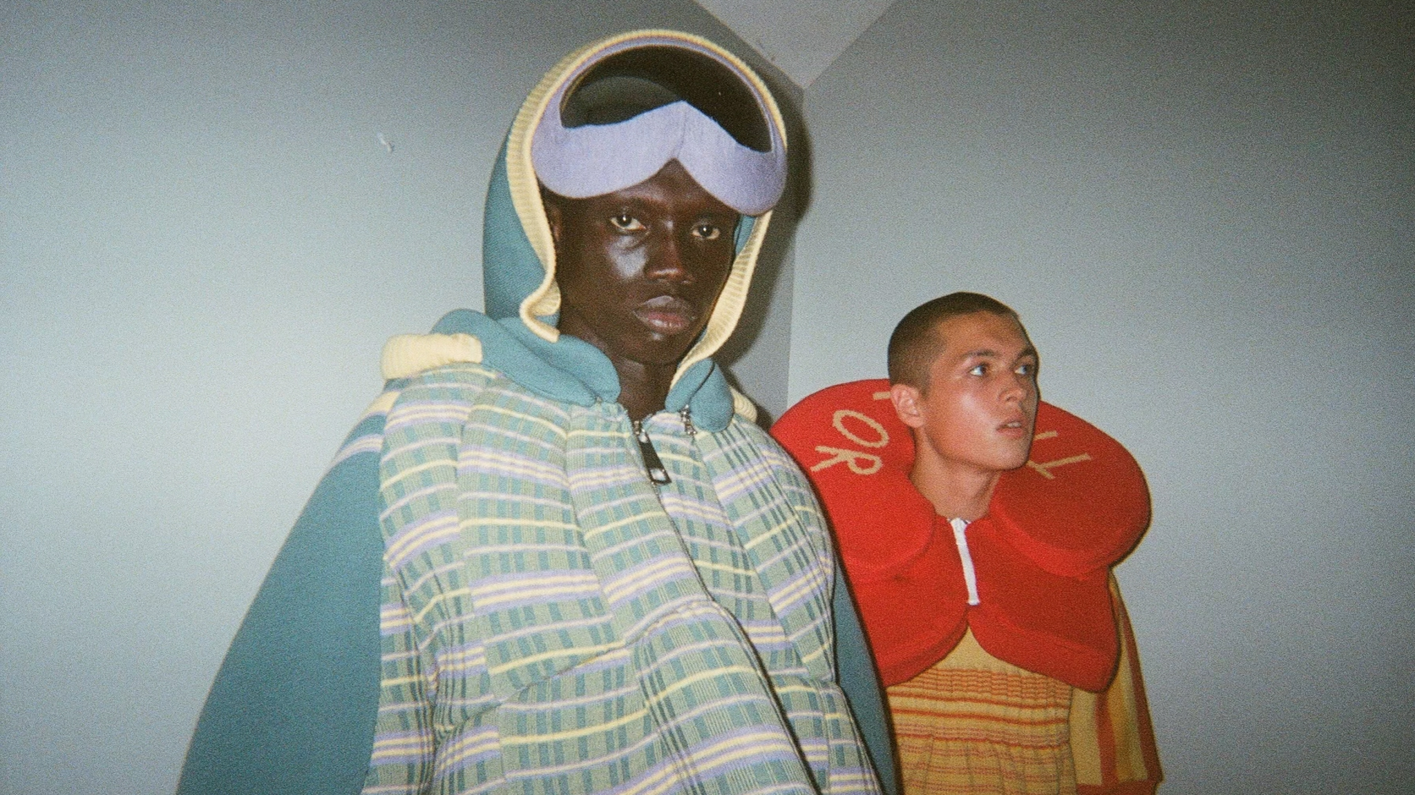 Two models stand backstage, one wearing a pastel green and blue plaid oversized knit puffer with ski goggles atop their head, and the other dressed in a bright yellow and red outfit, with a large red neck accessory resembling a life preserver.