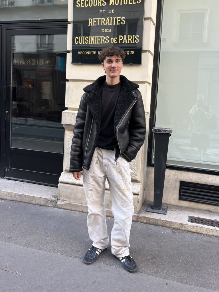 A student wears a black shearling coat, black sweater, white paint-splattered pants, and black adidas sneakers while standing in front of a brown and black building.