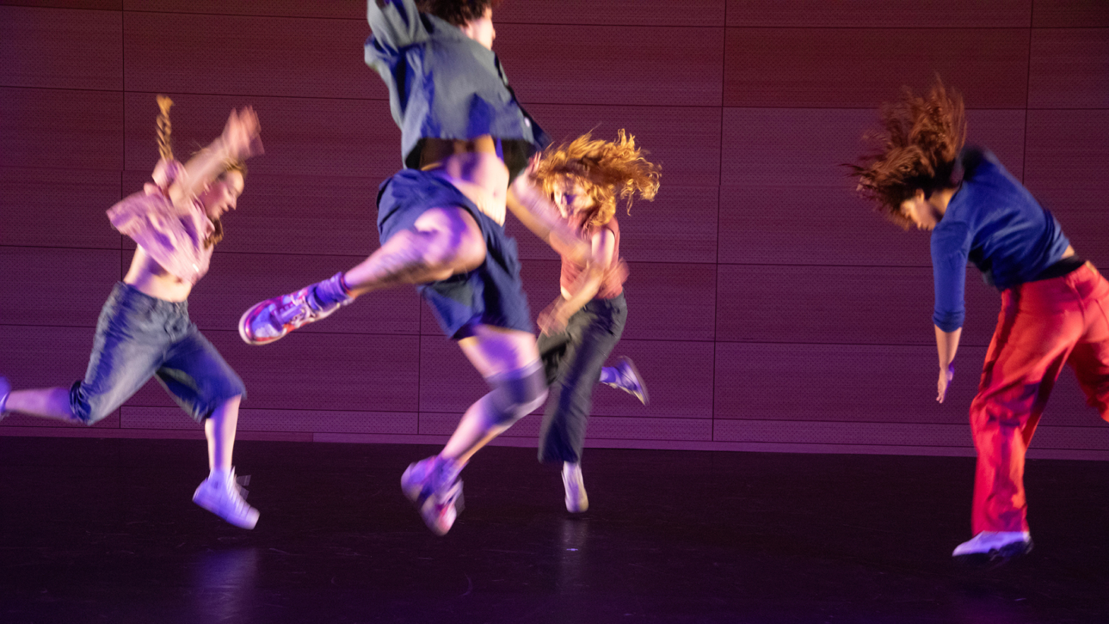 Four dancers mid-movement / jumping on stage, wearing colorful clothes