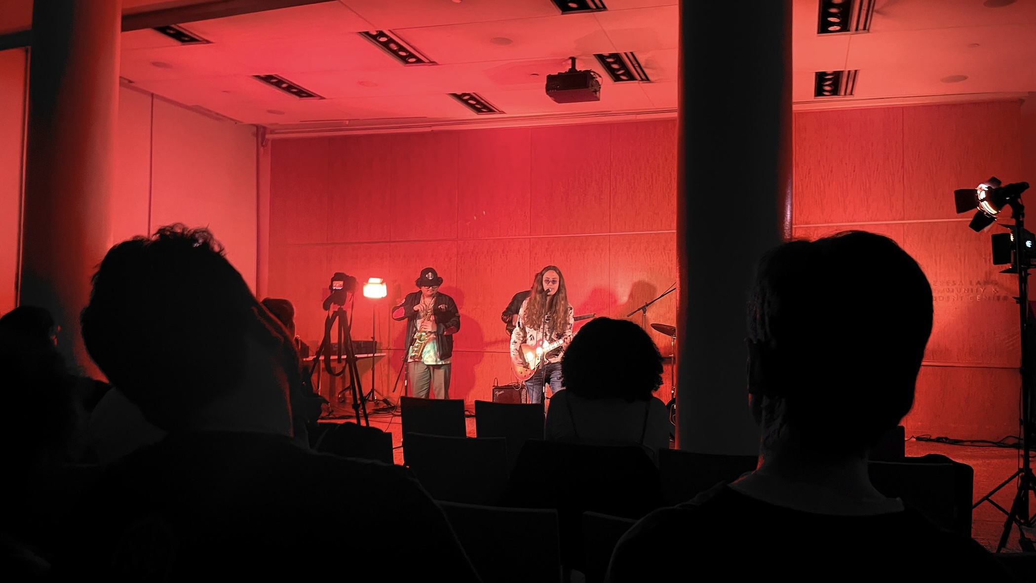 Three boys, one with long hair perform on red-lit stage strumming guitars and singing into microphone