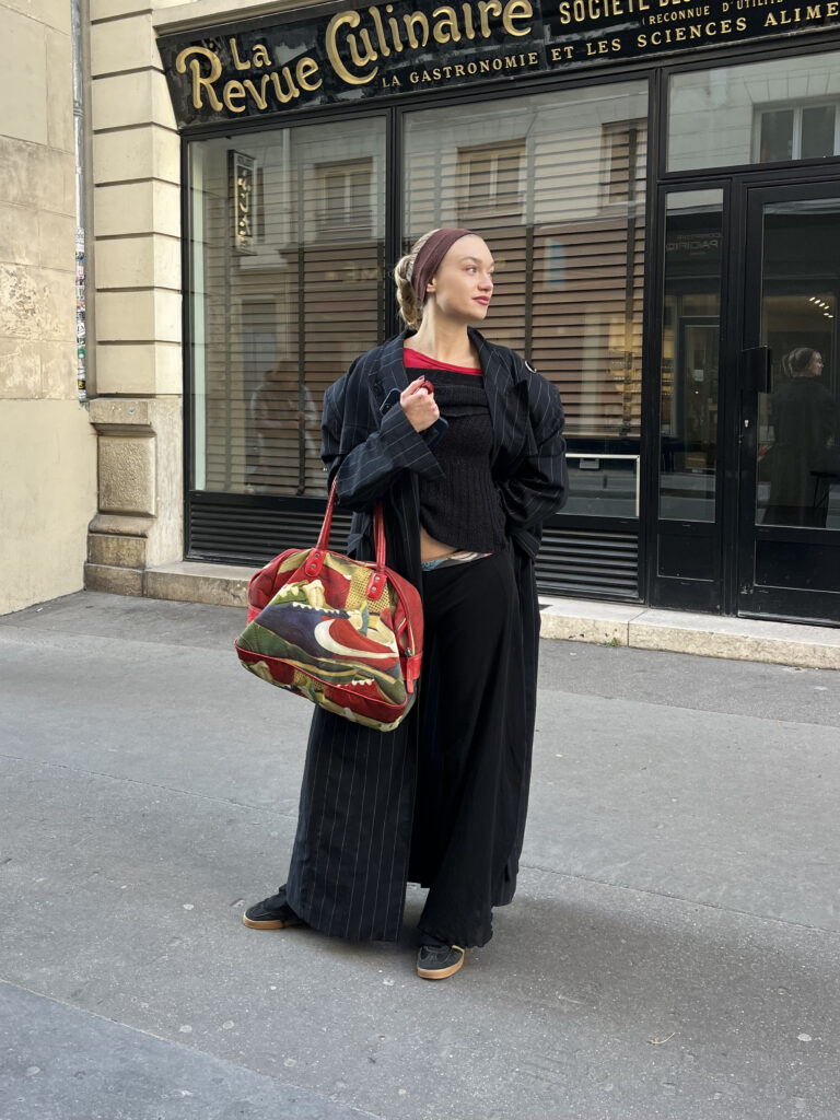 A student wears a brown headband, pinstripe trench coat, red T-shirt, black sweater, black trousers, black sneakers, and a colorful red bag while standing in front of a black and beige building.