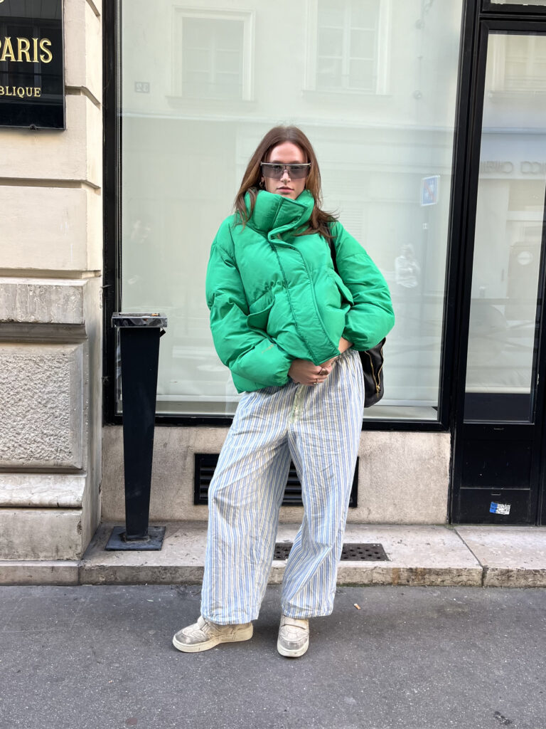  A student wears gray sunglasses, green puffer, blue and white striped pants, and white sneakers while standing in front of a beige and black building.