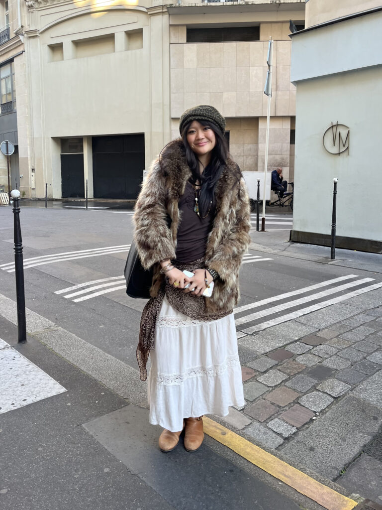 A student wears a plaid cap, brown fur coat, brown top, leopard scarf, white skirt, and brown boots while standing in front of brown and blue buildings.