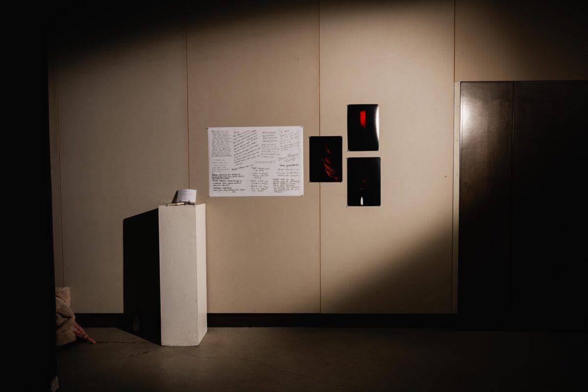 Three darker photographs and a larger white paper with written messafes on a wall next to a white stand with a notebook on top.