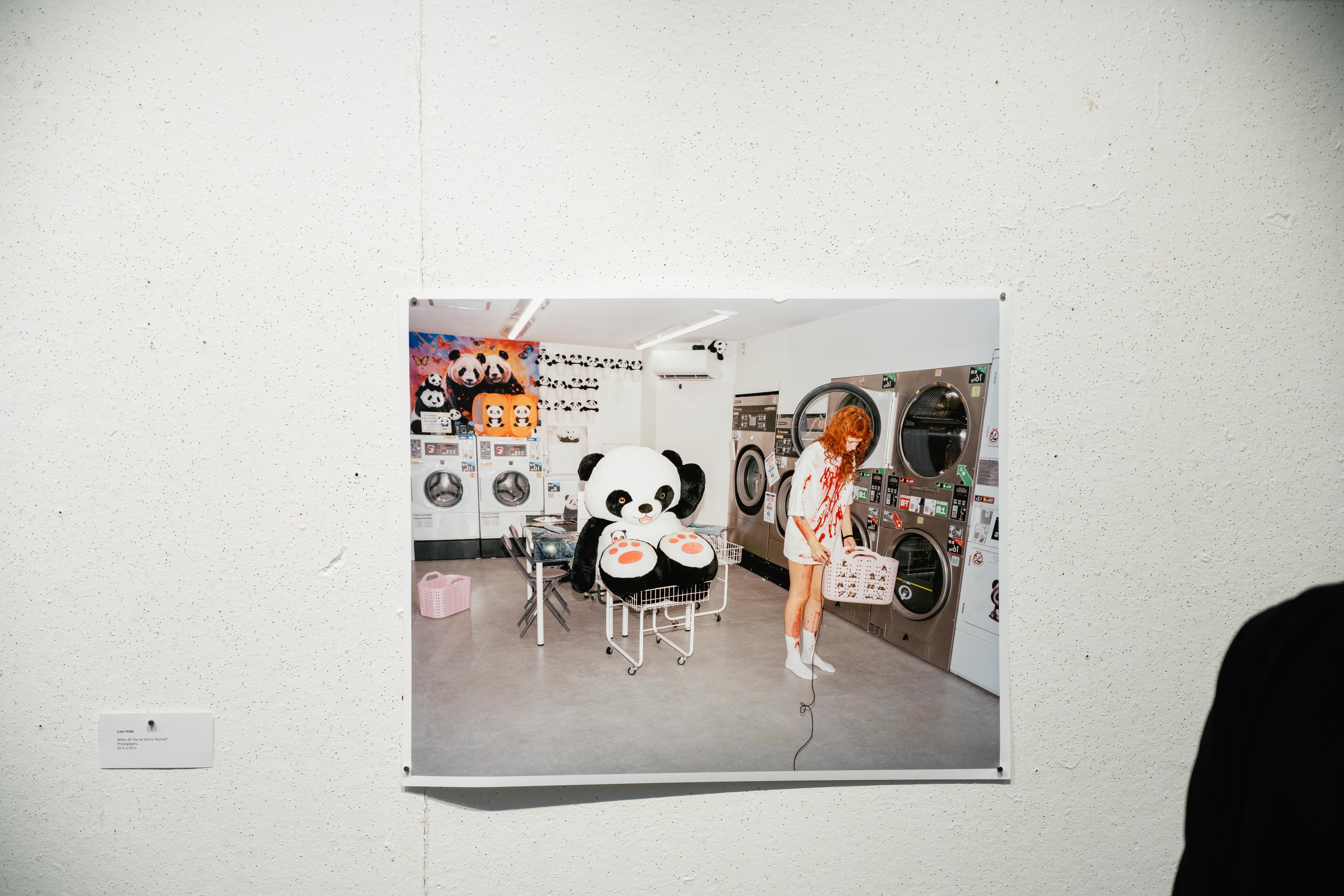 A photograph of a girl with a bloody t-shirt and a large stuffed animla panda in a landromat against on a white wall