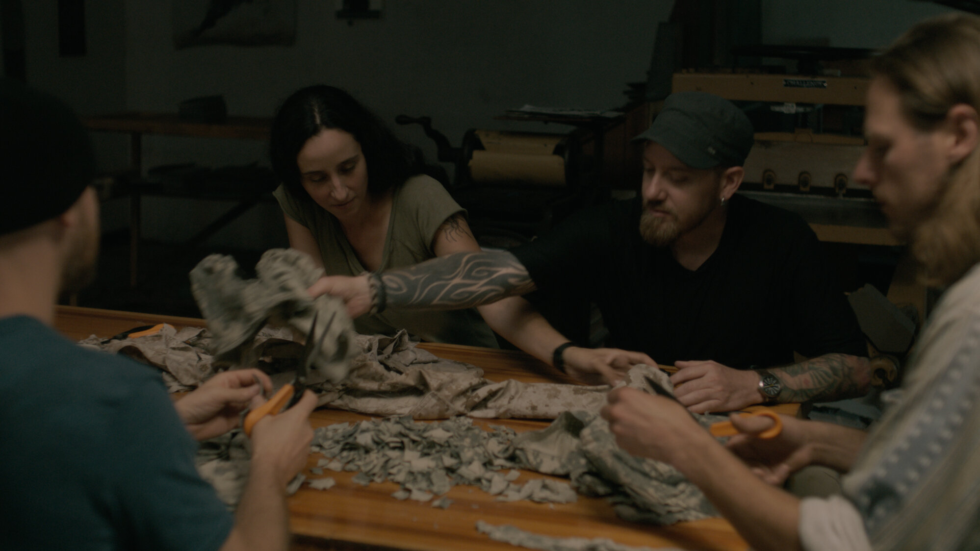 Two people, one in profile and one facing away from the camera, using scissors to cut up fabric, sit at a wooden table covered in scraps of military uniforms. Two people on the opposite side of the table, facing the camera reach across each other for different pieces of fabric.