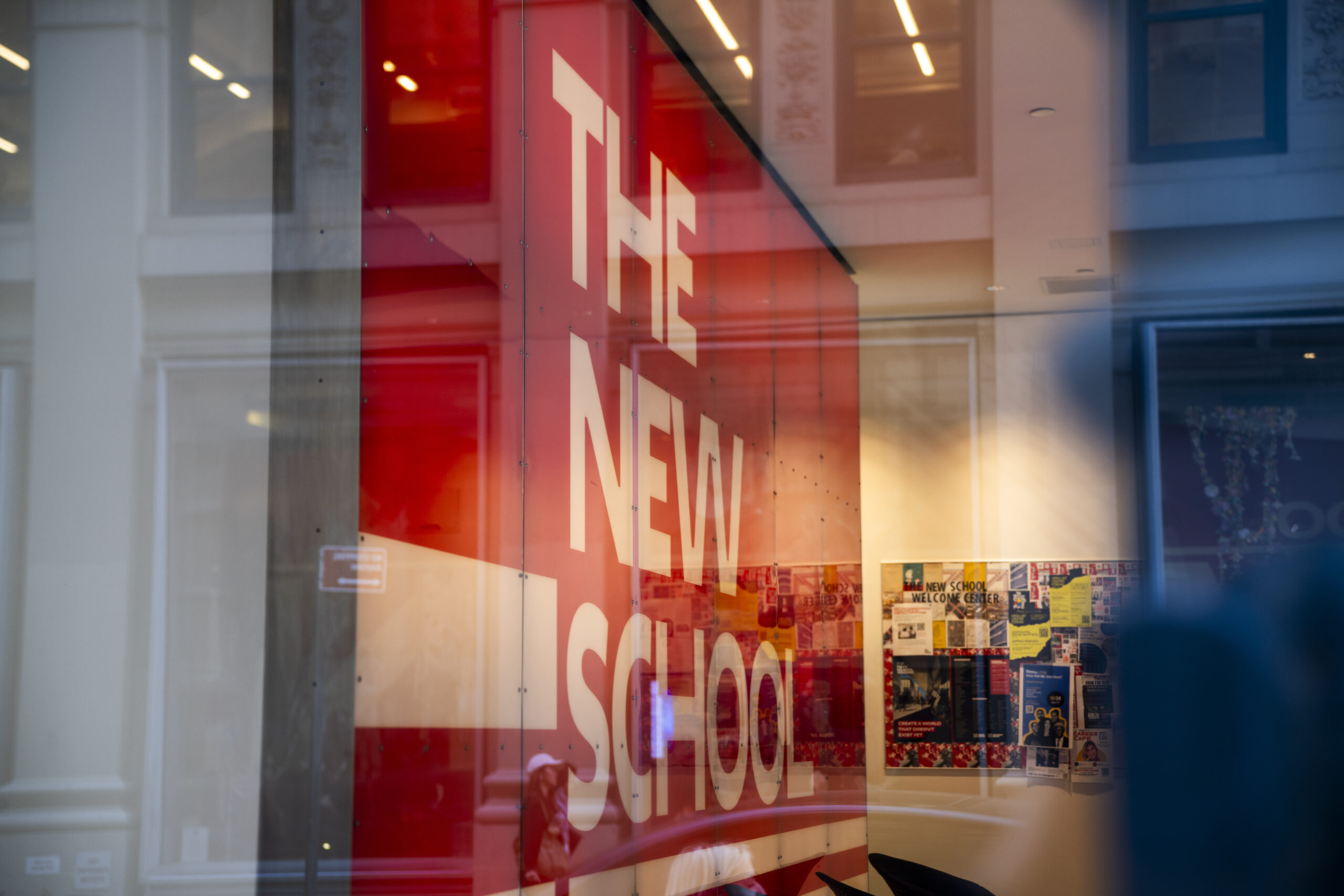 The New School logo in the Welcome Center. Words read “The New School” in white letters against a red wall. A bulletin board is on the right of the wall.