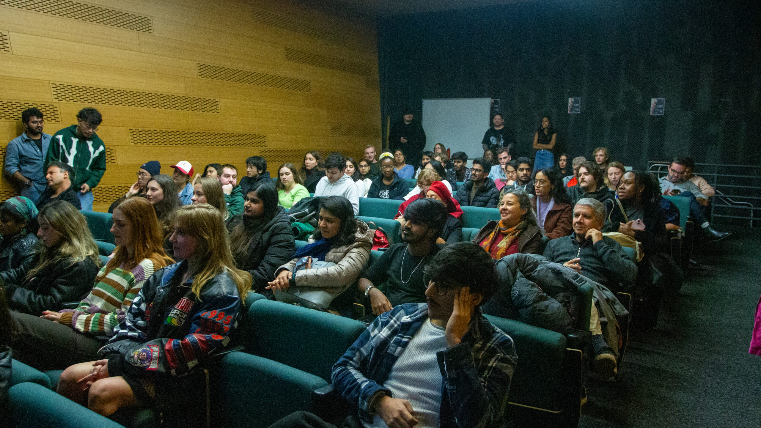 An audience sits in several rows of green chairs in a small auditorium.