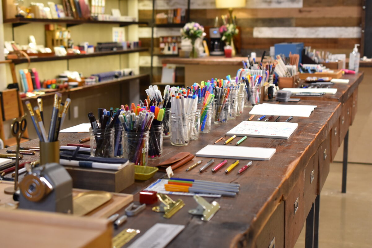 Pens in cups and pads of paper lined up on long wooden table inside Yoseka Stationery.