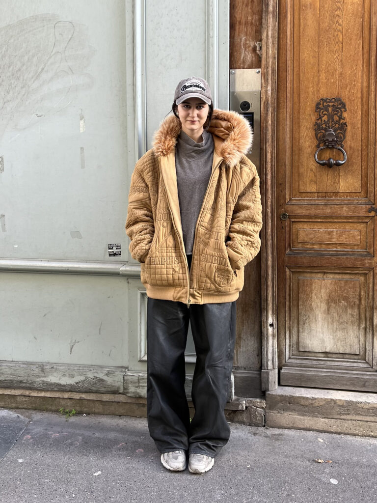 A student wears an orange coat, gray turtleneck, black jeans, white sneakers, and gray baseball hat while standing in front of a blue and brown building.