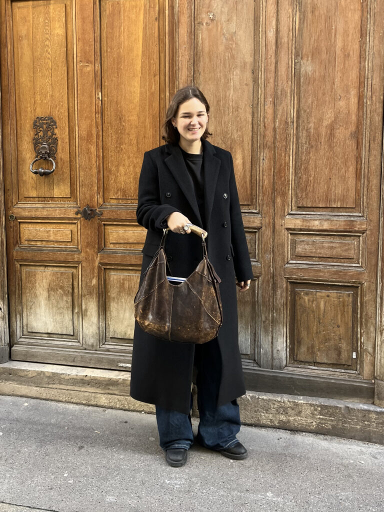 A student wears a black trench coat, black sweater, black jeans, black sneakers, and a brown bag while standing in front of a brown building.