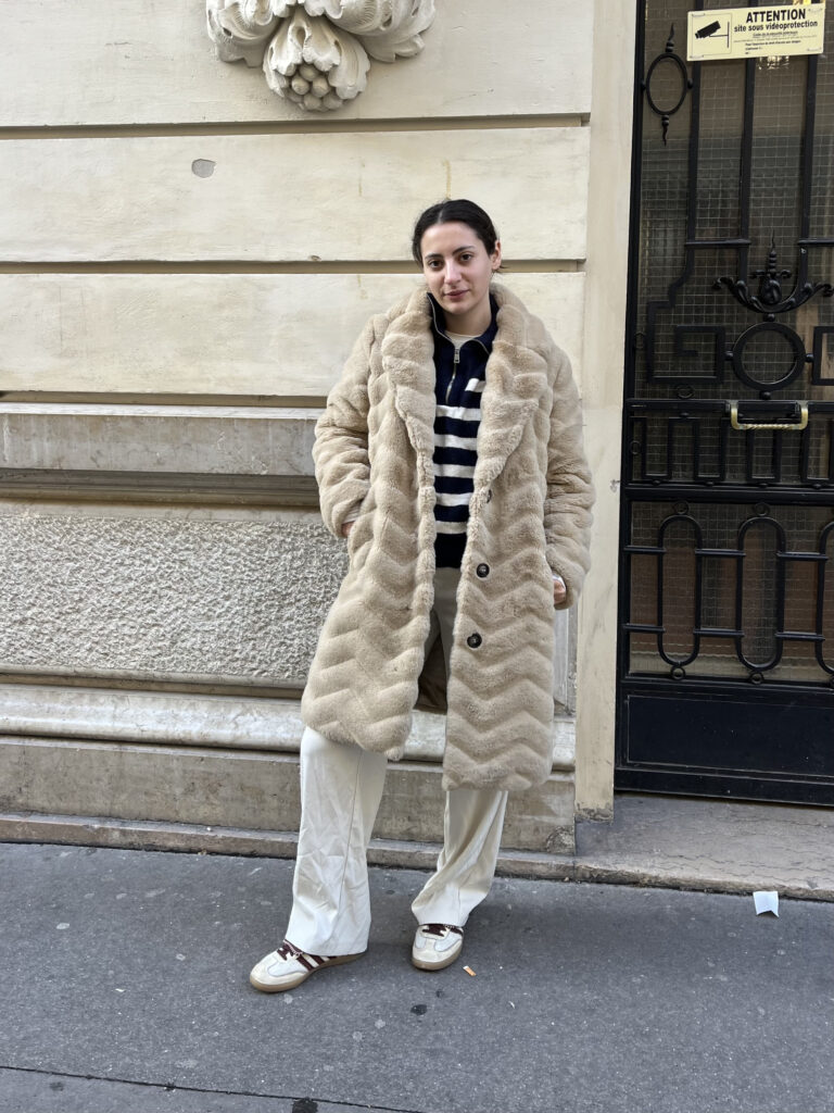 A student wears a beige coat, blue and white striped sweater, white pants, and white sneakers while standing in front of a beige building.