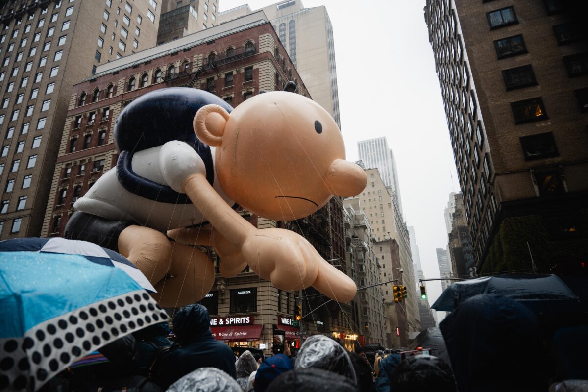 Large balloon of Greg Heffley in front of buildings against a gray sky, float down 6th Avenue.