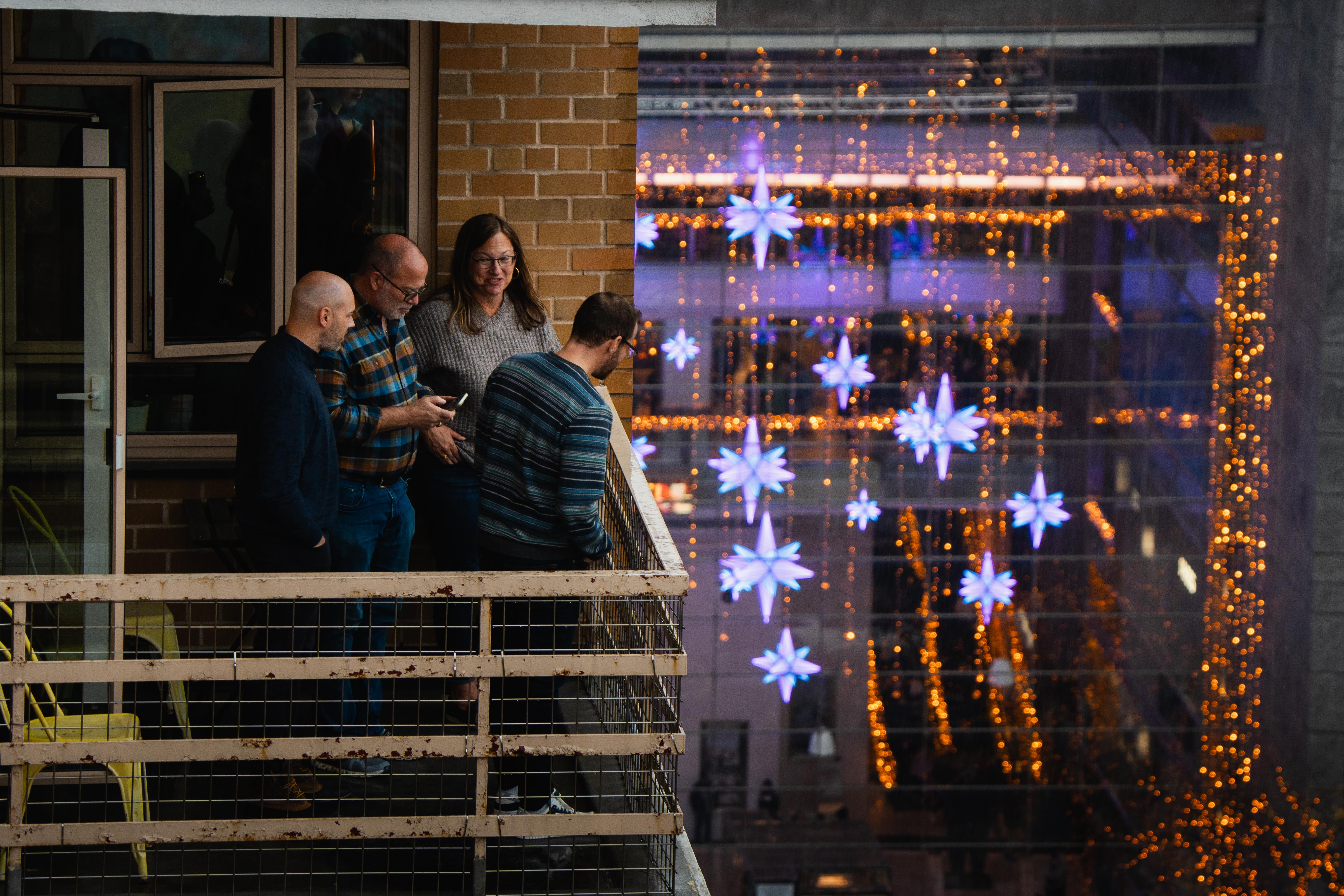 A group of four people in sweaters on a balcony look down towards the parade. In the background, large glass windows are covered in lights and lit-up snowflakes.