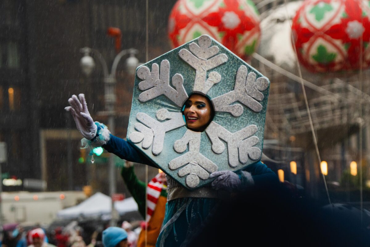 A person in a blue and white snowflake costume, with their head in the center, waves to the camera's left. In the background, Christmas ornament balloons float. 