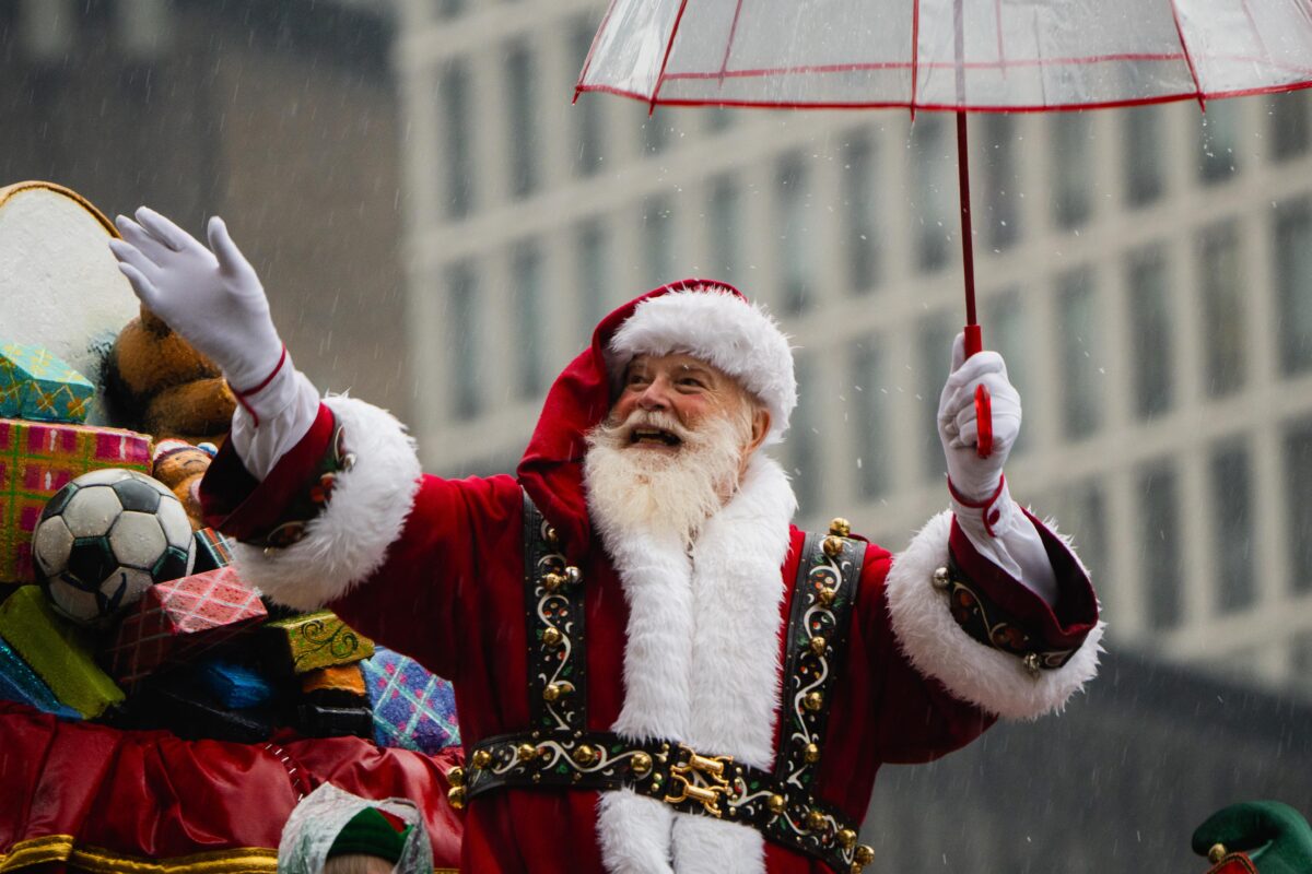 A man in a Santa costume, with a pile of presents in the background, holding a red-trimmed clear umbrella, waves to the left of the camera.