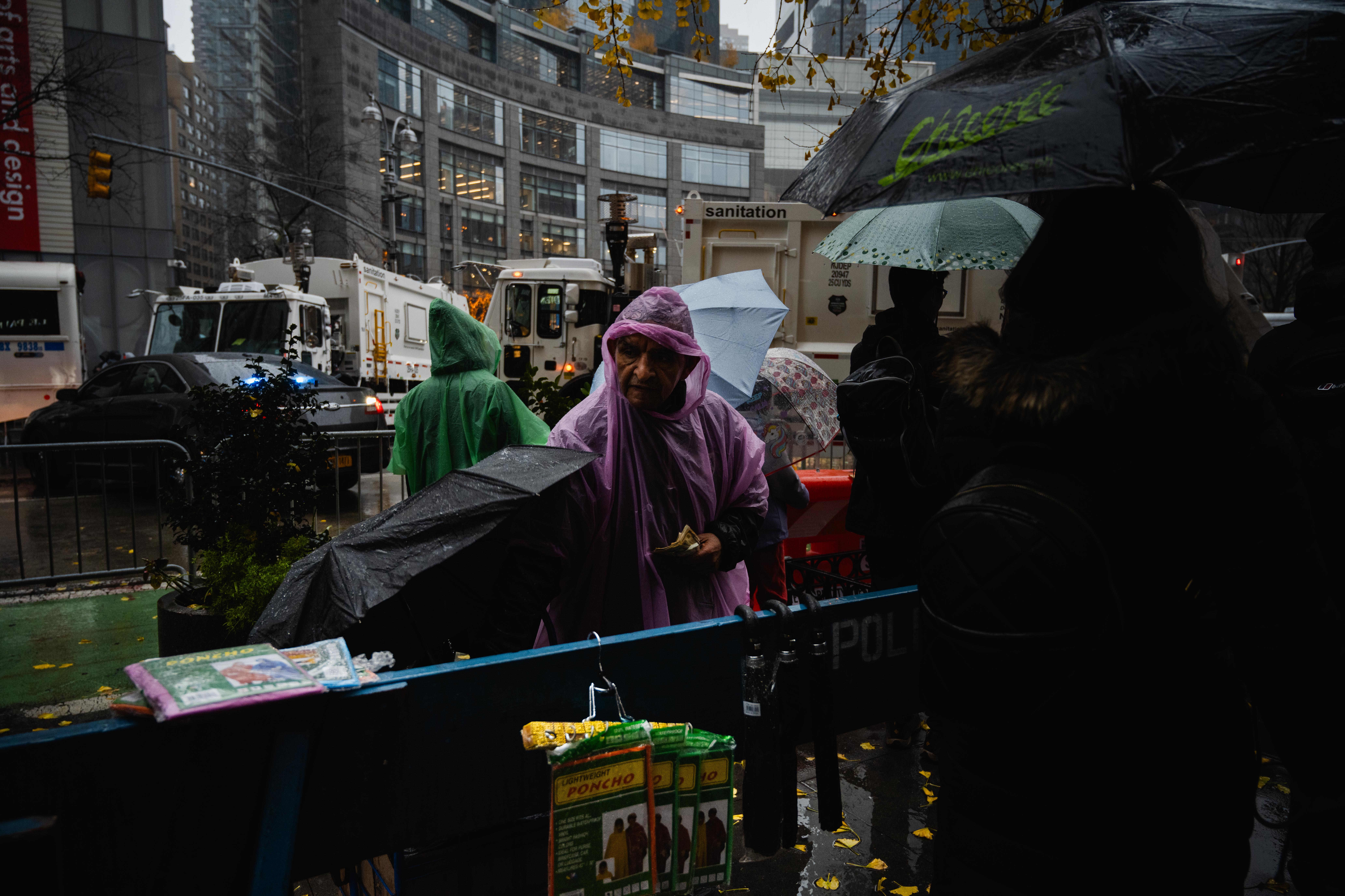 A man wearing a pink poncho looks down at ponchos in packages hanging off of a police barricade. In the background people in ponchos and umbrellas congregate.