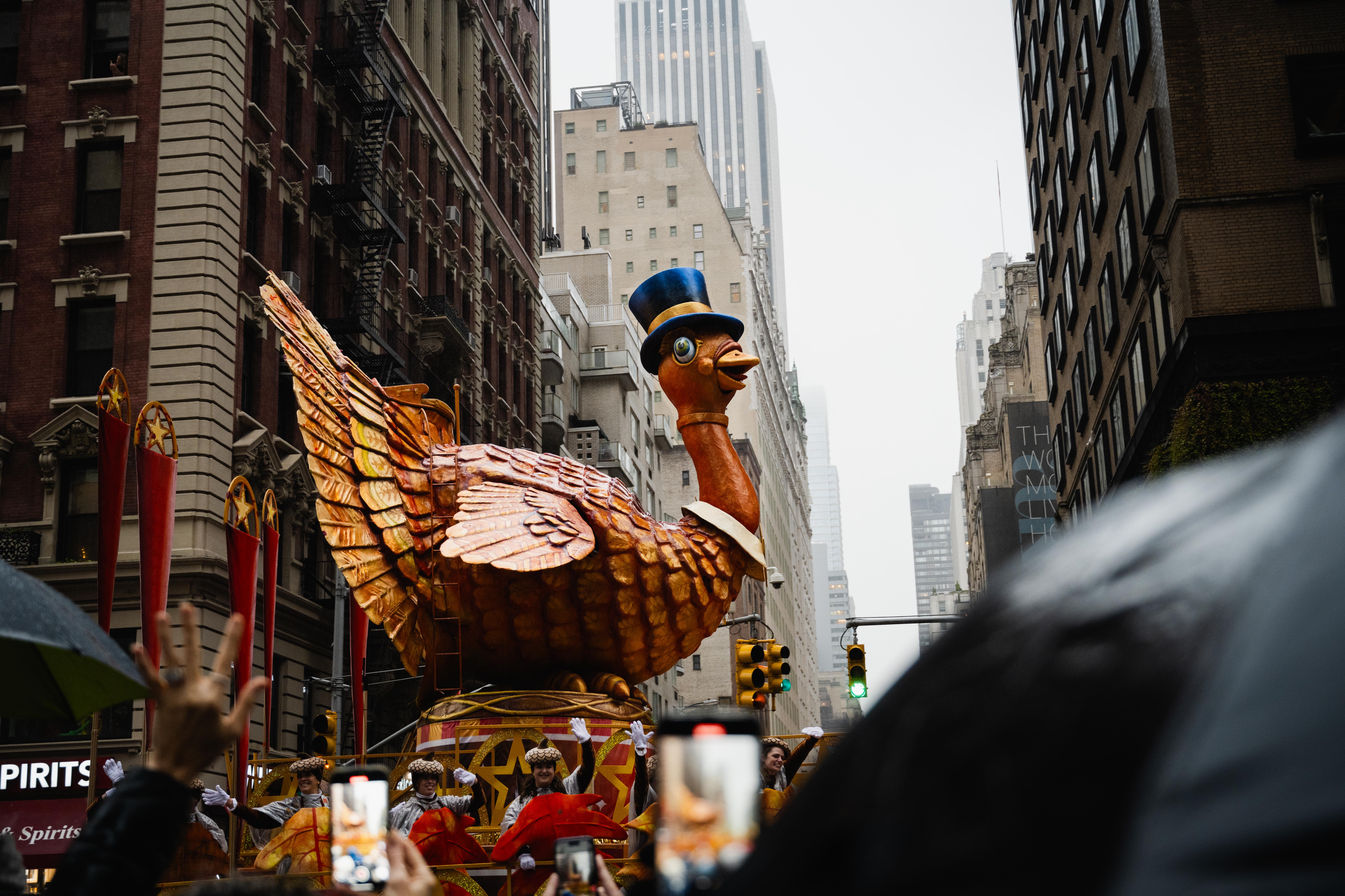 A large turkey float, wearing a top hat, floats in front of buildings and gray sky. In the foreground, people under umbrellas hold up their phones.