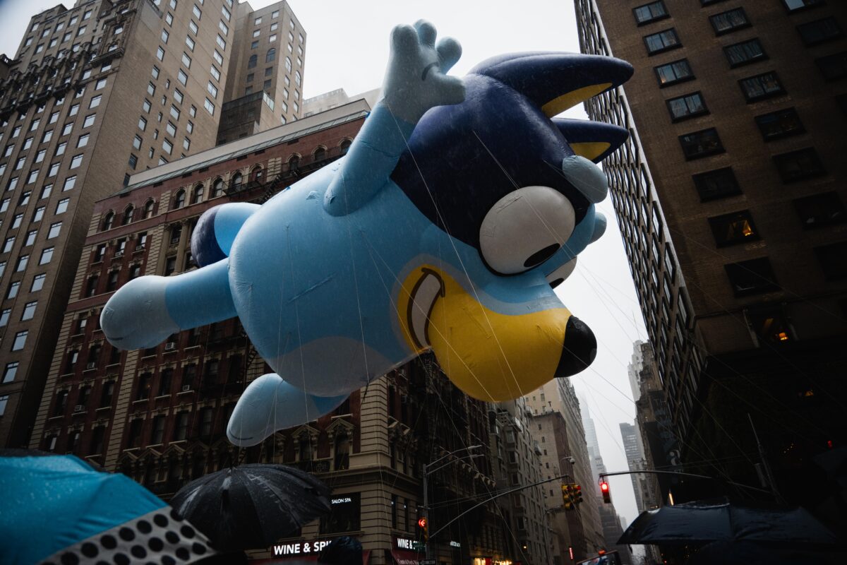 Large balloon of Bluey in front of buildings against a gray sky, float down 6th Avenue.