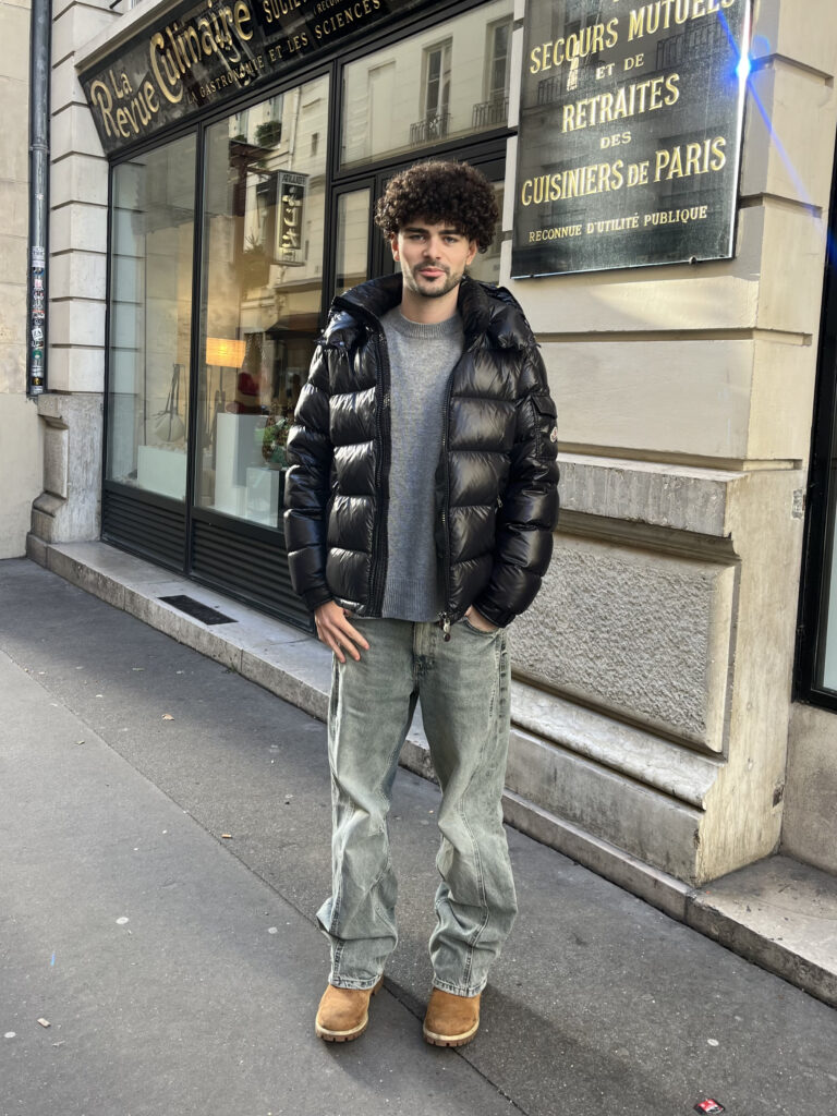 A student wears a black puffer coat, gray sweater, gray jeans, and brown boots while standing in front of a beige and black building.