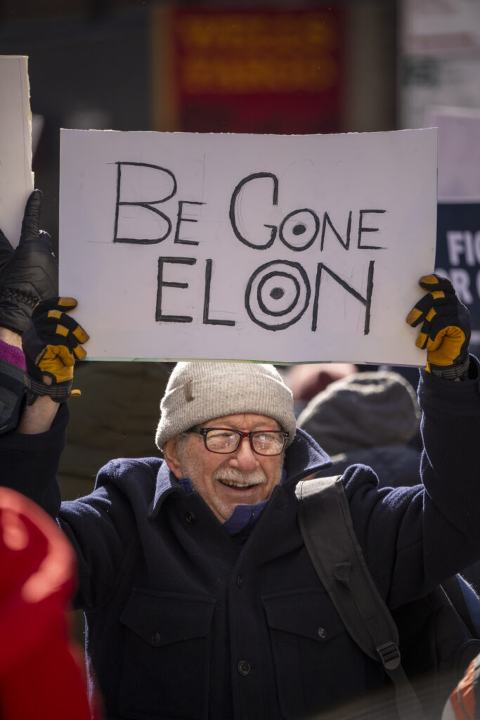 Protester holds a sign reading “Be Gone Elon”. 