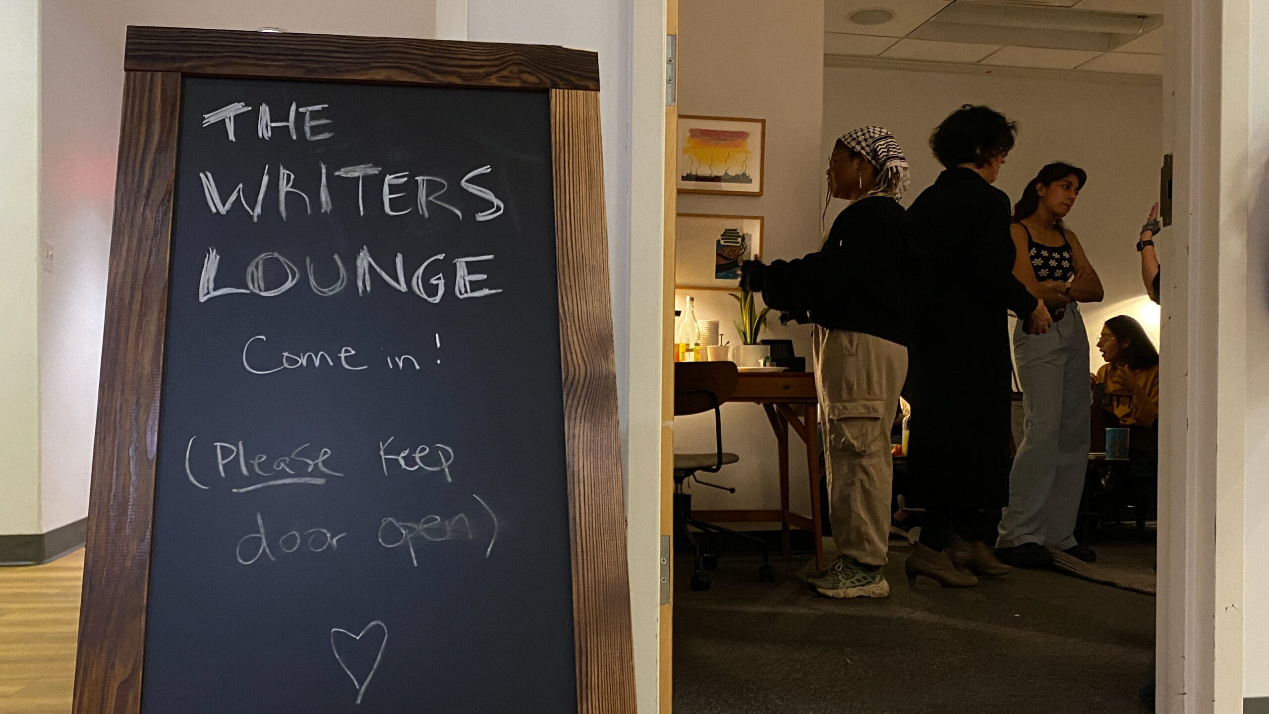 A folding wooden sign that reads “The Writers Lounge. Come in! (Please keep door open),” with a heart beneath, written in chalk. The sign rests near the open door of the lounge, where three people stand inside talking.