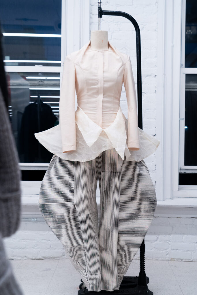 A mannequin wears a structured cream jacket with a flared peplum and oversized striped pants with wide, sculptural legs.