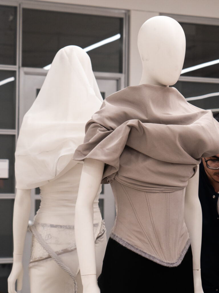 Two mannequins display garments, one in a draped gray corset with a black skirt, another a white dress with a sheer veil.